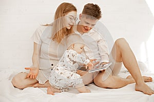 Portrait of young smiling family in white clothes with plump baby infant toddler sitting on white cotton bed cloths.