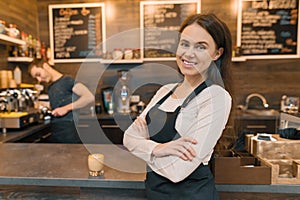 Portrait of young smiling female cafe worker, standing at the counter. Woman with folded hands, professional baristas team cafe