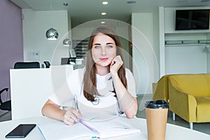 Portrait of young smiling female doctor making notes in notebook while sitting behind the table with phone and coffee inside of