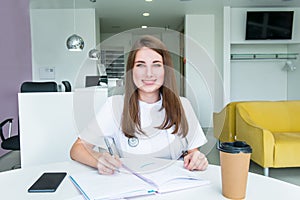 Portrait of young smiling female doctor making notes in notebook while sitting behind the table with phone and coffee inside of