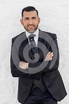 Portrait of young smiling handsome man groom fiance with short dark hair wearing black suit tuxedo, standing at wall.