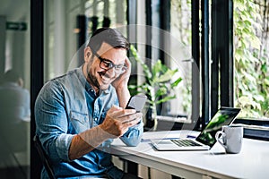 Portrait of young smiling happy handsome successful businessman entrepreneur freelancer working from office using smart phone