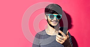 Portrait of young smiling hipster looking in smartphone, wearing cyan sunglasses and gray shirt, on pink coral background.