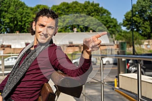 Portrait of a young smiling Latin man sitting on a plaza bench and pointing to the side