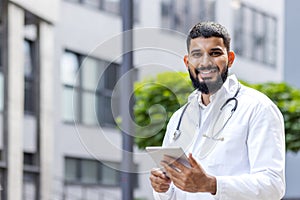 Portrait of a young smiling male Muslim doctor standing in a white coat and with a stethoscope outside a clinic and