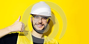 Portrait of young smiling man, builder engineer, showing thumbs up, wearing white construction safety helmet, glasses and jacket.