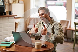 Portrait of young smiling man, online tutor shows thumbs up at laptop, video chats with someone who did good job, guy