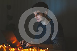 Portrait of young smiling man using laptop in dark room at home with garlands.