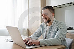 Portrait of young smiling man using laptop sitting at desk, writing in notebook. Cheerful guy browsing internet