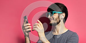 Portrait of young smiling man using smartphone and wireless earphones, wearing cyan sunglasses, on pink coral background.