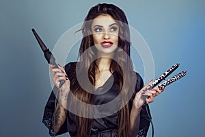 Portrait of young smiling model with perfect teeth and long hair wearing silk peignoir holding curling wand and hair straightener