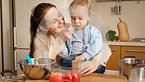 Portrait of young smiling mother feeding her baby son with fruit puree on kitchen. Concept of little chef, children cooking food,