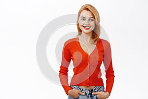 Portrait of young smiling redhead woman, looking inspired and hopeful, standing over white studio background