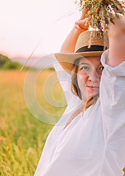 Portrait of young smiling woman dressed in light summer clothes and straw hat walking by high green grass meadow with wildflowers