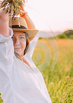 Portrait of young smiling woman dressed in light summer clothes and straw hat walking by high green grass meadow with wildflowers