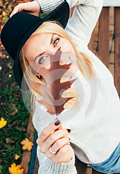 Portrait of a young smiling woman holding oak leaf in hand agains her face, wearing white sweater and black felt hat