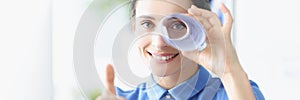 Portrait of a young smiling woman holds thumbs up and looks into pipe made of paper