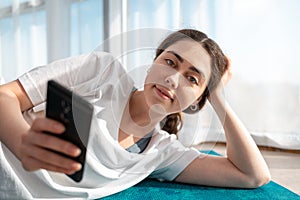 Portrait of young smiling woman lies on the sports mat and uses a smartphone. Windows in the background. Communication and network