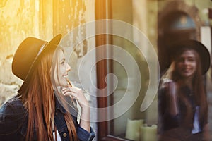 Portrait of young smiling woman looking at the shop window. Model wearing stylish wide-brimmed black hat. City lifestyle