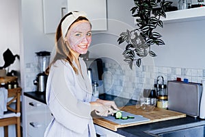 Portrait of young smiling woman with pink clay facial mask cutting cucumber for salad on the kitchen. Every day home