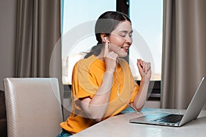 Portrait of a young smiling woman sitting at a desk wearing headphones chatting online on a laptop. The concept of online courses