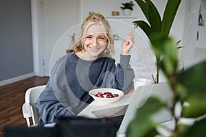 Portrait of young smiling woman, sitting in room, watching videos on laptop, eating lunch or breakfast, healthy meal
