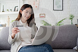 Portrait of young smiling woman sitting on a sofa at home. She is holding a credit card and using a computer to shop online