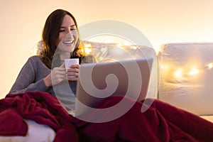 Portrait of young smiling woman sitting on sofa with laptop and cup of coffee or tea and looking on the screen