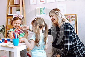 Portrait of young smiling woman sitting at table around children playing. Teacher stretching hand with paper butterfly.