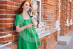 Portrait of young smiling woman talking on mobile phone standing leaning on old red stone wall at city street in summer