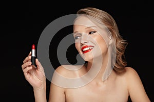 Portrait of young smiling woman with wavy hair holding red lipstick in hand joyfully looking aside over black background