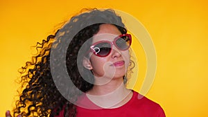 Portrait of young spanish girl with curls puts on sunglasses on yellow background. Tempting woman smiling to camera