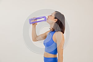 Portrait of young sporty muscular woman with dark hair wear blue sportswear, drinking water from purple plastic bottle.
