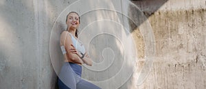 Portrait of young sporty woman in sportswear leaning agaist concrete wall in the city.