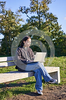 Portrait of young student, girl using laptop, sitting in park on bench, typing on computer