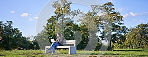 Portrait of young student, girl using laptop, sitting in park on bench, typing on computer