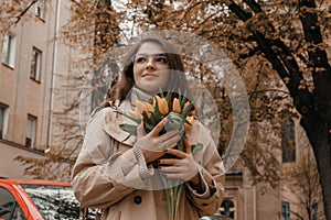 Portrait of young stylish attractive woman walking in city, street fashion, autumn trend, coat, holding flowers bouquet