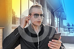 Portrait of a young stylish guy of Caucasian appearance of a businessman in sunglasses, jacket and shirt, adjusts his headphones