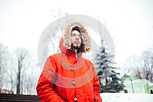 Portrait of a young stylish man with beard dressed in red winter jacket with hood and fur on his head stands against the backdrop