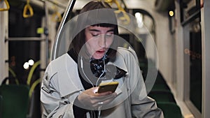 Portrait of young stylish woman in headphones listening to music and browsing on mobile phone in public transport. Young