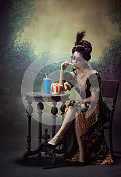 Portrait of young stylish woman, medieval royal person in elegant dress eating fries with soda against dark vintage