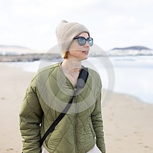 Portrait of young stylish woman wearing green padded jacket, hoodie, wool cap and sunglasses on long sandy beach in