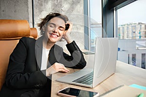 Portrait of young successful business woman in suit, sitting in office with laptop, smiling and looking happy, enjoys