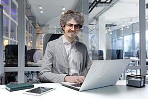 Portrait of young successful businessman financier, man inside office at workplace sitting using laptop smiling and