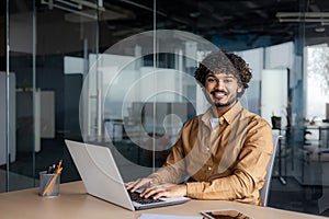 Portrait of young successful Indian man at workplace inside office, businessman smiling and looking at camera, man at