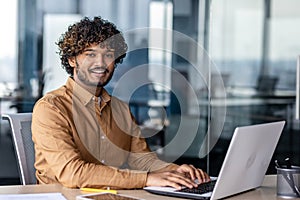Portrait of young successful Indian man at workplace inside office, businessman smiling and looking at camera, man at work using