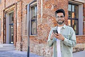 Portrait of young successful student outside university campus, man smiling looking at camera and using app on phone