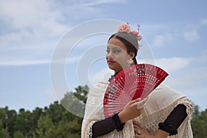 Portrait of young teenage girl in black dance dress, white shawl and pink carnations in her hair, dancing flamenco with a red fan