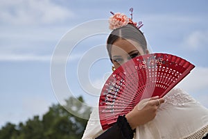 Portrait of young teenage girl in black dance dress, white shawl and pink carnations in her hair, dancing flamenco with a red fan