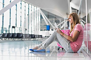 Portrait of young teenage girl sitting on the floor and using digital tablet while waiting for her flight in airport